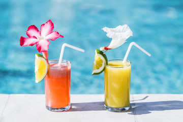Two glasses of fresh cold fruit juice on poolside. With straw, slice of lime and tropical flower