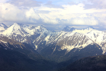 Fototapeta na wymiar View from top of mountain peak. Mountain valley in blue haze and snowy mountain range. Ravine and cloudy sky. 
