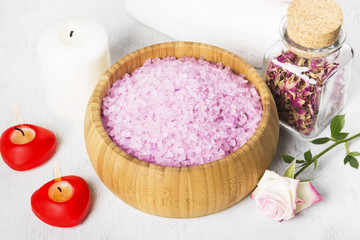 Obraz na płótnie Canvas Bath salt with aroma of a rose in a wooden bowl, petals and a fresh pink rose, towels and candles on a white background