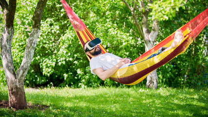 Obraz na płótnie Canvas Young bearded man wearing virtual reality goggles relaxing in a garden hammock. Lifestyle VR fun and relax concept
