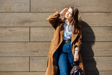 Young stylish brunette woman student in brown coat, blue jeans posing over wooden texture wall in sunny day.