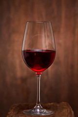 Close-up of crystal half-ful wineglass with red wine standing on wooden stand against wooden background.