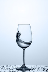 Splashing transparent water wave in the wine glass while standing on the glass against light background.