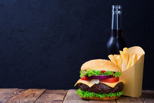 Burger with french fries and drink on rustic wooden table with black chalkboard background as copy space