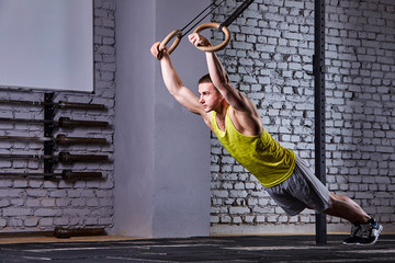 Young athlete man working out in gym pull ups with gymnastic rings against brick wall in the cross fit gym.