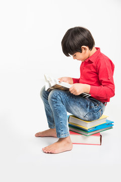 Indian Little Boy Or Kid Sitting On Stack Of Books While Reading A Book. Isolated Over White