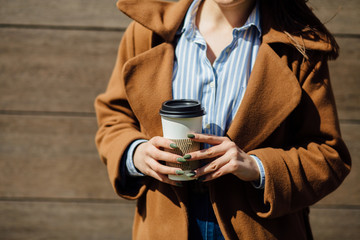 A cropped photo of disposable cup with coffee in hand of stylish woman. A beautiful model look woman standing with a cup of coffee in hands beside the wooden wall background.