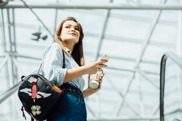 sale, consumerism and people concept - beautiful young woman in striped shirt, blue jeans and bagpack holding mobile phone and disposable cup of coffee and rising on escalator in shopping center.