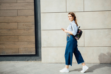 Full height photo of brunette woman student in striped shirt, blue jeans and backpack walking beside the concrete and wooden wall background.