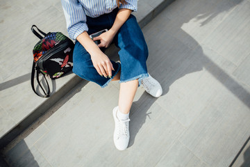 Unrecognizable hipster woman with bagpack and smartphone sitting on stairs in city, wearing a striped shirt, blue jeans and white sneakers. From above