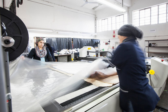 Female factory workers unrolling textile onto pattern cutting machine in clothing factory