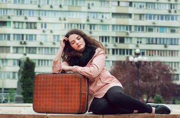Romantic girl with retro style suitcase sitting outdoor and waiting for transport on big city street.