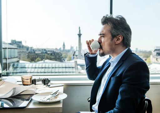Businessman drinking coffee by restaurant window with rooftops views, London, UK