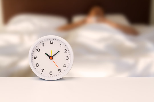 White alarm clock on white desk on blurred man sleeping on white bedding sheet, time concept