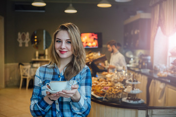 Young blonde woman enjoying coffee or chocolate in cozy cafe. Female holding cup of cappuccino. Blurred interior behind the model.