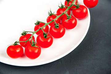 Long branch of organic ripe fresh cherry tomatoes on a white plate laying on black texture background. Healthy vegetarian food.