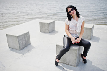 Portrait of brunette girl listening music at headphones from mobile phone, wear on women's leather pants and white blouse, sunglasses, against stone cubes at beach of lake.