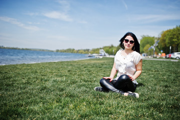 Naklejka na ściany i meble Brunette girl on women's leather pants and white blouse, sunglasses, sitting on geen grass against beach of lake.