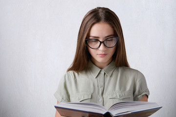 A young female student with long straight hair wearing shirt and eyeglasses holding a book reading something. A young girl reading encyclopedia isolated over white background. Educational concept.