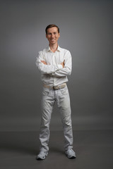 Young smiling man in white shirt and jeans stands on a gray background with arms crossed on chest.