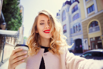 Young girl in a glass of coffee in her hand makes selfie on the background of a sunny street of the old city