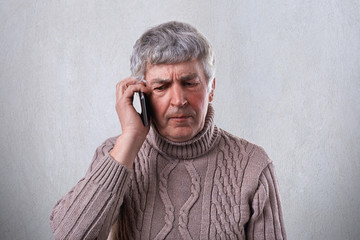 An angry mature man with gray hair and wrinkles holding smartphone in his hand communicating with his children. Old man being unsatisfied while talking over the mobile phone.