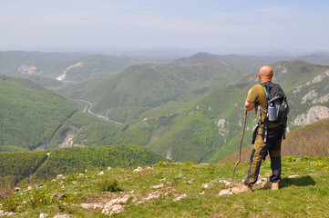 Fototapeta na wymiar Hiker with backpack relies on a stick and enjoy the view. Man on top of the mountain look at the valley below