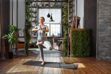 Young woman in homeware practicing balance yoga pose on carpet in her comfy bedroom.