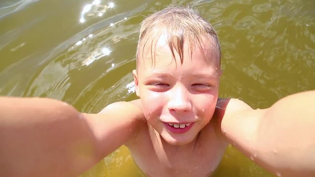Closeup portrait of cute funny kid taking selfie on summer beach. Little boy uses modern technology. Face and body wet from river water. Child sitis in water and poses for camera then swims away.