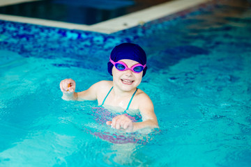 Portrait funny little girl in blue cap and pink goggles playing in swim pool. Close up