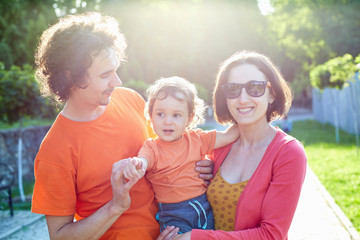 Family with child walks and plays in city park. A young father, mother and a small son dressed in orange t-shirt. The child funny dresses up sunglasses and plays with them. Child in the hands of his
