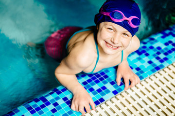 Portrait funny little girl in blue cap and pink goggles playing in swim pool. Close up