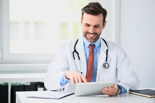 Happy doctor sitting at desk, using tablet