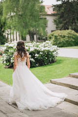 Fototapeta premium The delicate beautiful bride walks in the park in a wedding dress with lace and plume, bush with flowers. Fine art