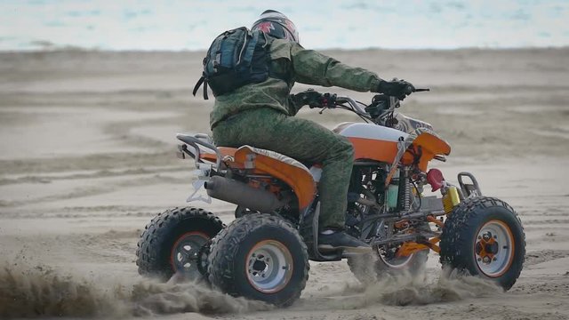 A man wearing a helmet on his head performs an extreme trick on a sports ATV on the beach area, the sand flies out from under the wheels of the vehicle