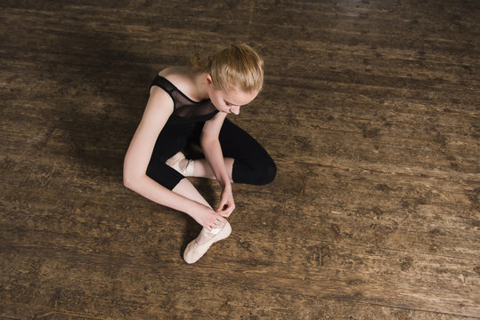 Young ballerina or dancer girl putting on her ballet shoes on the wooden floor. Female dancer ties on her pink ballet slippers with ribbons. Top view.