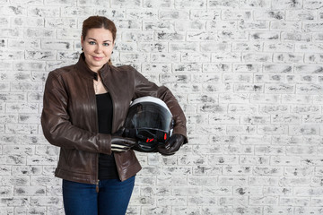 Fototapeta na wymiar Young woman in leather protective clothes holding motorcycle helmet in hands, copyspace with brick wall of garage
