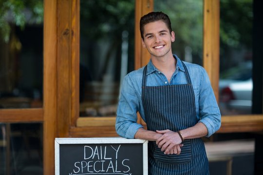 Smiling waiter leaning on menu board outside the cafe - Powered by Adobe