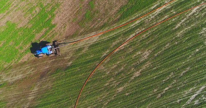 Tractor fertilizing field with liquid manure pumped from distant tanker truck via giant hose, aerial view.