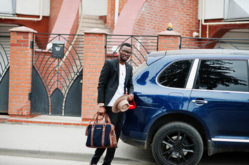 Stylish black man at glasses with hat, wear on suit with handbag against luxury car. Rich african american businessman.