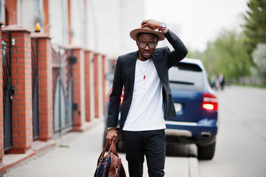 Stylish black man at glasses with hat, wear on suit with handbag against luxury car. Rich african american businessman.