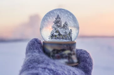 Fototapeten Girl in knitted mittens holding glass ball with firtrees, house and artificial snow falling inside the ball with winter landscape at the  background © yos_moes