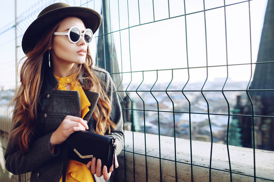Outdoor waist up portrait of young beautiful woman with long hair. Model wearing stylish hat, sunglasses, clothes, holding small bag. City lifestyle. Female fashion concept. Copy, empty space for text