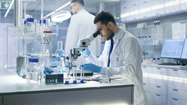 Research Scientist Adjusts Sample in a Petri Dish with Surgical Pincers and Looks on it Under Microscope. He's Working in a Modern Laboratory.