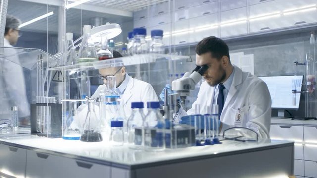 In a Busy Modern Laboratory Two Scientists Conduct Experiments. Chief Research Scientist Dictates the Results He Sees in a Microscope to His Assistant. Shot on RED EPIC-W 8K Helium Cinema Camera.