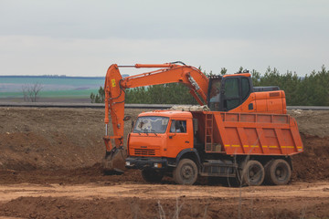Heavy excavator loading dumper truck on road construction among fields