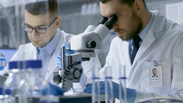 In a Modern Laboratory Two Scientists Conduct Experiments. Chief Research Scientist Dictates the Results He Sees in a Microscope to His Assistant. Shot on RED EPIC-W 8K Helium Cinema Camera.