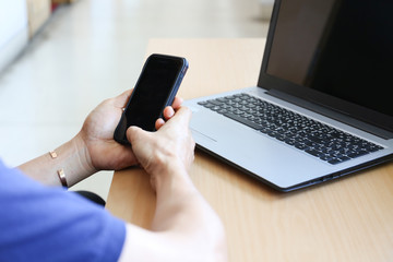 Man is holding mobile phone black display on hands in front of laptop on wooden desk