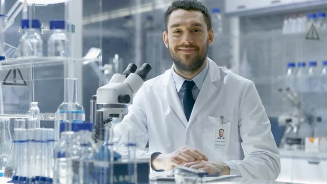 Young Male Research Looks at Sample under Microscope then Looks into Camera and Smiles. He's Sitting in a High-End Modern Laboratory with Beakers, Microscope and Working Monitors Surround Him. 