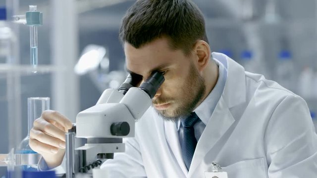 Research Scientist Looks into  Microscope. He's Working in a High-End Modern Laboratory with Beakers, Glassware, Microscope and Working Monitors Surround Him. 